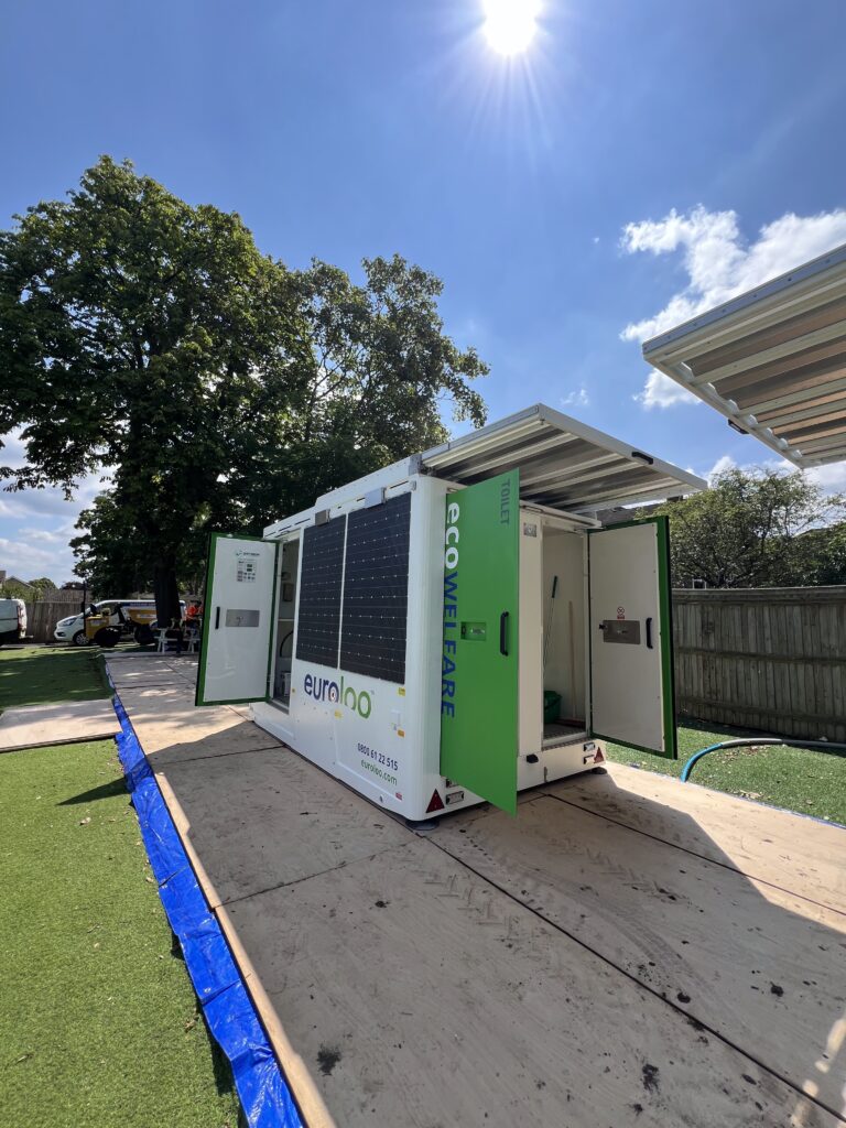 A Euroloo Ecowelfare Unit With Solar Panels In Use At A Mid-Sized Construction Or Industrial Site, Surrounded By Cars And Fencing