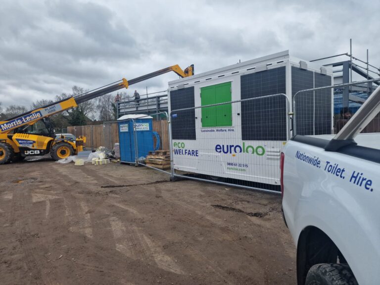 A 12Ft Euroloo Ecowelfare Unit With Solar Panels In Use On A Live Construction Or Maintenance Site, Positioned On A Tarmac Surface Near Green Space And Wooden Fencing.