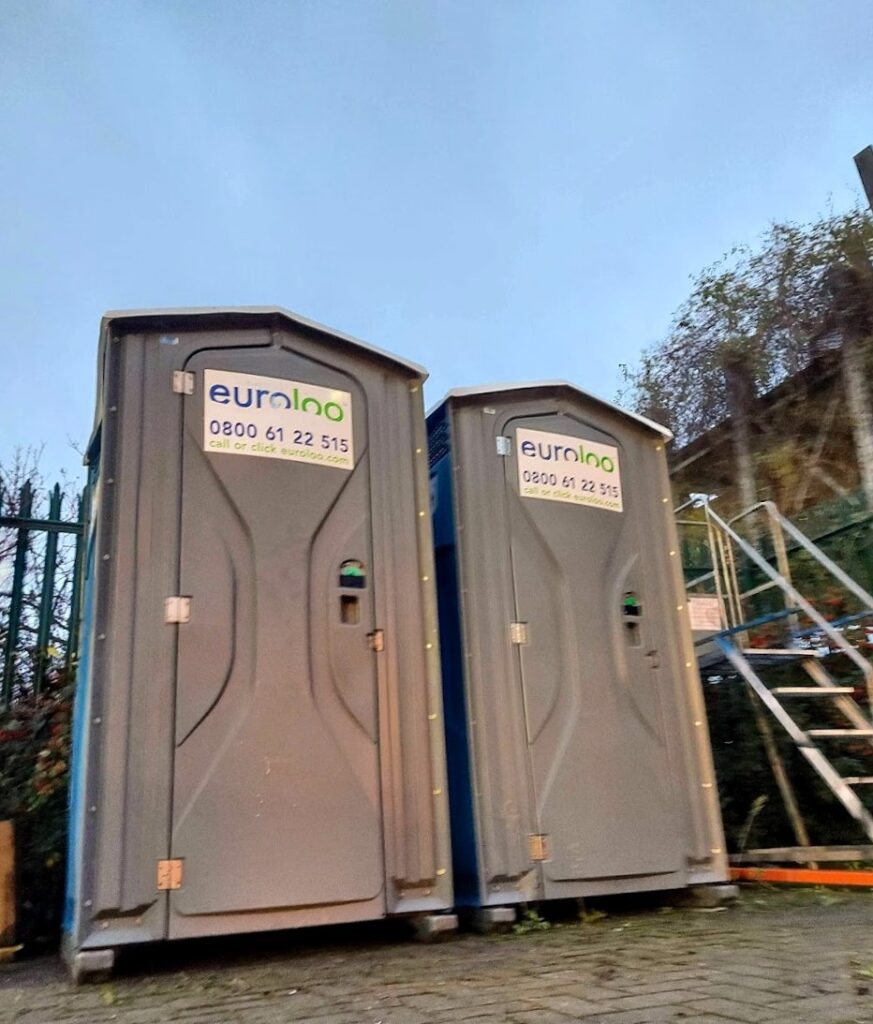 Two Clean Portable Toilets Positioned On A Uk Business Site, Representing Workplace Toilet Provision For Staff, Visitors And Delivery Drivers.
